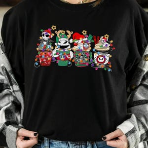 May include: Black crew neck t-shirt featuring four cartoon coffee cups decorated with Christmas lights and characters from The Nightmare Before Christmas. The cups have different designs and colors, including Jack Skellington and Zero.