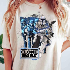 May include: Cream-colored t-shirt featuring a graphic of Clone Troopers from "Star Wars: The Clone Wars." The design includes blue and white armored figures with weapons, set against a textured background. The text "THE CLONE WARS" is displayed in a black and blue box.