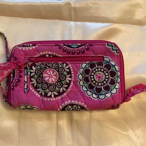 May include: A quilted pink wallet with a floral pattern in white, teal, and purple. The wallet has a zipper closure and a wrist strap. The floral design is circular and repeated across the surface of the wallet.