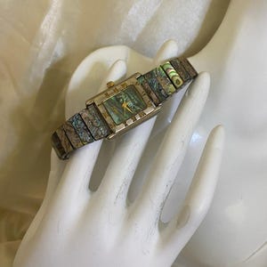 May include: A rectangular wristwatch featuring a square face with an iridescent abalone shell dial and gold-tone hands. The watch band is composed of linked segments inlaid with matching iridescent abalone shell, creating a cohesive look.