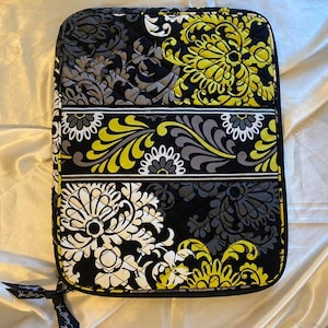 Könnte beinhalten: Eine gesteppte, rechteckige Hülle mit einem Blumenmuster in Schwarz, Grau, Weiß und Gelb. Das Design zeigt große stilisierte Blumen und wirbelnde Blätter. Ein schwarzer Reißverschluss und ein schwarzes Band mit Text sind sichtbar.