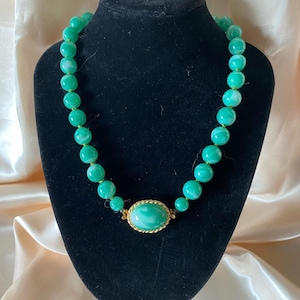Puede incluir: Un collar de cuentas verdes con un colgante ovalado dorado. El collar se exhibe en un maniquí negro. Las cuentas son de un color verde vibrante con vetas blancas. El colgante es de un color verde similar con un marco dorado.