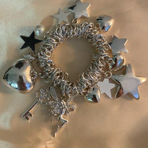 May include: Silver charm bracelet featuring a coiled band and various charms. Charms include stars, hearts, a fairy, a key, and a pearl. The bracelet is displayed on a satin surface.