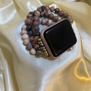 May include: A smartwatch with a rose gold casing and a black screen, featuring a multi-strand bracelet made of round, variegated stone beads in shades of dusty pink, mauve, and charcoal gray, connected by small silver-toned metal spacer beads.