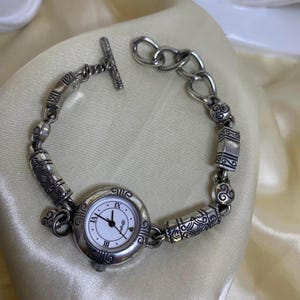 May include: Silver-toned watch bracelet with a white watch face featuring Roman numerals. The bracelet has decorative links and a toggle clasp. The watch face has black hands and a second hand.