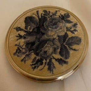 May include: A round, vintage compact with a gold-tone metal frame. The top features a detailed needlepoint design of dark blue roses and leaves on a tan background. The compact is closed and appears to be in good condition.
