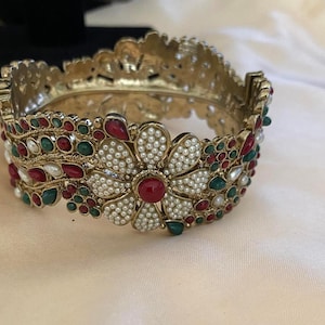 May include: A gold-toned bangle bracelet with intricate floral and geometric designs. The bracelet is adorned with red, green, and white faux gemstones and pearls. The central flower design features a large red gemstone center.