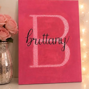 May include: A pink canvas with a large, white, glitter-filled letter "B" and the name "Brittany" in cursive script. The canvas is a vibrant pink, and the text is centered. The background includes a vase of pink roses and string lights.