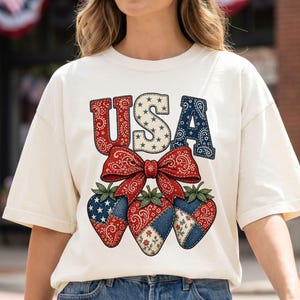 May include: Cream-coloured t-shirt featuring a patriotic design. The word "USA" is displayed in red, white, and blue, with a bow and strawberry graphic. The strawberries have red, white, and blue patterns.