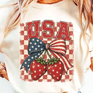 May include: A cream-coloured t-shirt with a patriotic design. The graphic features the word "USA" in red, a blue bow with white stars, red and white striped ribbons, and two strawberries with white stars, set against a red and white chequered background.