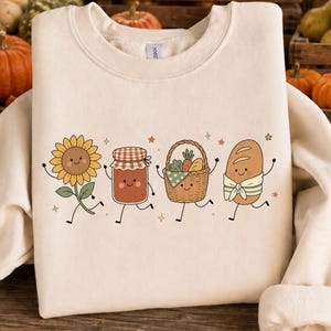 Op de afbeelding: Een crèmekleurig sweatshirt met ronde hals en een grafische print van vier antropomorfe figuren: een lachende zonnebloem, een jampot met een geruit deksel, een gevlochten mand vol groenten en een brood dat een sjaal draagt.