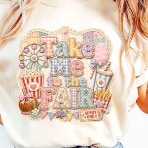 May include: Cream-colored t-shirt featuring a colorful fairground graphic. The design includes the text "Take Me to the Fair" in patchwork letters, along with illustrations of a Ferris wheel, popcorn, cotton candy, and other fair treats.