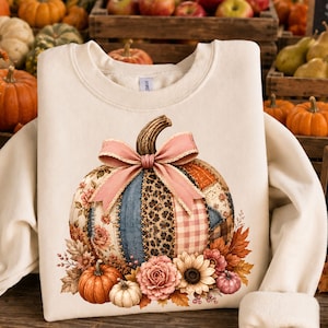 Peut inclure: Un sweat-shirt couleur crème arborant un motif central de citrouille en patchwork ornée d'un nœud en ruban rose. Le design de la citrouille inclut des motifs floraux, léopard et à carreaux, entouré d'illustrations de petites citrouilles et fleurs.