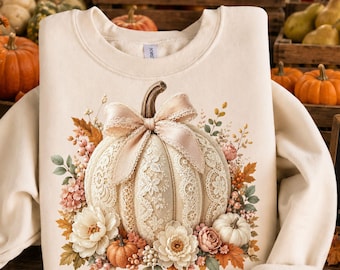 May include: Cream-colored sweatshirt featuring a detailed illustration of a white pumpkin adorned with lace, a pink bow, and surrounded by autumn flowers and leaves. The design evokes a fall or harvest theme.