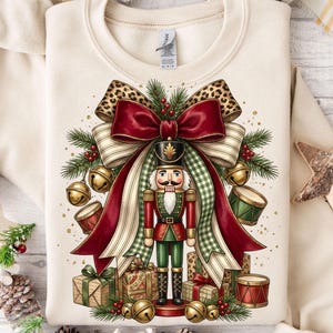 May include: A cream-colored crewneck sweatshirt featuring a graphic of a nutcracker standing in front of a large red and green bow. The design includes pine branches, gold bells, small drums, and wrapped gift boxes with leopard print accents.