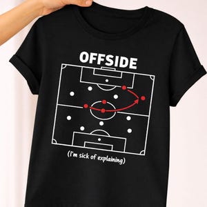 Op de afbeelding: Zwart T-shirt met het woord "OFFSIDE" boven een voetbalveld diagram. Het diagram toont een spel met rode stippen en een gebogen pijl. De tekst "(I'm sick of explaining)" staat eronder.