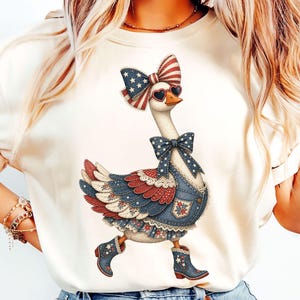 Puede incluir: Camiseta color crema con una ilustración de un ganso patriótico. El ganso lleva un lazo, gafas de sol y un peto vaquero con detalles en rojo, blanco y azul. El ganso también lleva botas.