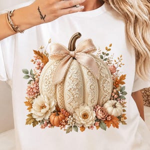 Floral lace pumpkin PNG displayed on a clean white t-shirt mockup. The fall design includes a large cream pumpkin with lace border detailing, elegant floral lace panels, a soft blush bow, ivory flowers, pink blossoms, orange fall leaves, berries, sage greenery, and mini pumpkin accents. Great for coquette fall shirt designs, lace pumpkin PNG listings, pumpkin bow PNG art, autumn PNG design files, pumpkin patch shirts, boutique fall graphics, and trendy fall sublimation projects.