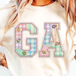Op de afbeelding: Crèmekleurig T-shirt met de letters "GA" in een patchwork-ontwerp. De letters zijn omlijnd in roze en versierd met bloemen- en kanten details. Het patchwork bevat pastelkleuren en ruitpatronen.