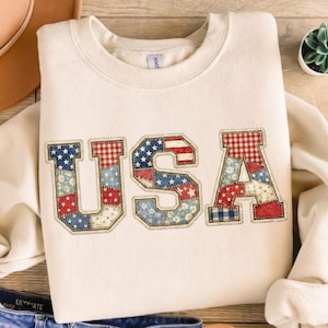 May include: Cream-colored sweatshirt with the word "USA" in a patchwork design. The letters are filled with red, white, and blue patterns, including stars and stripes, and outlined with a decorative stitch.