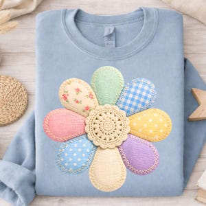 May include: A light blue sweatshirt featuring a large flower design. The flower petals are various colors and patterns, including floral, polka dots, and gingham. The center of the flower is a beige crochet doily.