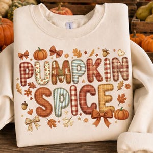 May include: Cream-colored sweatshirt with the words "PUMPKIN SPICE" in a patchwork design. The letters are decorated with pumpkins, leaves, and bows in shades of orange, brown, and green. A cozy fall-themed garment.