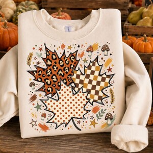 May include: A cream-coloured crewneck sweatshirt featuring a central graphic of three large maple leaves. One leaf has a leopard print, one a brown and white checkerboard pattern, and one a brown polka dot design, surrounded by small mushrooms and stars.