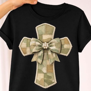 May include: Black t-shirt featuring a cross design. The cross is embellished with a camouflage pattern, a bow, and a pearl-like trim. The bow is centered with a cluster of pearls.