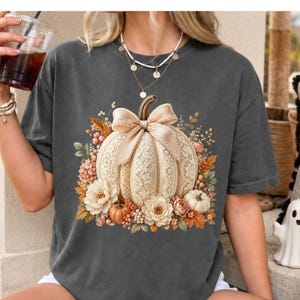 Boutique fall lace pumpkin PNG shown on a washed gray t-shirt mockup with a casual autumn lifestyle feel. The design features a cream lace pumpkin with delicate lace border accents, a blush bow, floral panels, ivory flowers, soft pink blossoms, fall leaves, berries, sage greenery, and mini pumpkins. Ideal for coquette fall PNG designs, pumpkin sweatshirt graphics, fall pumpkin shirt listings, floral pumpkin PNG files, lace border PNG projects, and trendy autumn tee mockups.