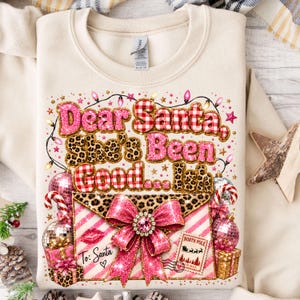 Pode incluir: Uma camisola de gola redonda cor de creme com uma impressão gráfica que diz Dear Santa, She's Been Good... Ish. O design inclui purpurinas cor-de-rosa, padrão xadrez vermelho e branco, estampado leopardo, um grande laço rosa e um envelope.