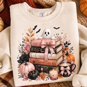 May include: A cream-coloured crewneck sweatshirt featuring a graphic print of a stack of five books topped by a small white ghost. The design includes a pink bow, black and pink pumpkins, a steaming mug with a pink bow, a candle, and bats.