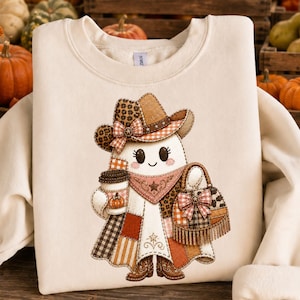 May include: A cream-colored crewneck sweatshirt featuring a central graphic of a ghost wearing a cowboy hat, a bandana, and boots. The ghost holds a coffee cup with a pumpkin print and a fringed handbag, all decorated in patchwork patterns.