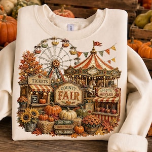 May include: A cream-colored sweatshirt featuring a graphic print of a county fair scene. The design includes a ferris wheel, a carousel, a ticket booth, and stalls for caramel apples and hot cider, surrounded by pumpkins, sunflowers, and autumn leaves.