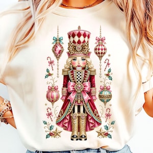 Könnte beinhalten: Ein cremefarbenes T-Shirt mit einer zentralen Grafik eines Nussknackers in einer rot-goldenen Uniform mit einem Hut mit Leopardenmuster, umgeben von hängendem Weihnachtsschmuck, Zuckerstangen, Stechpalmenblättern und goldenen Sternen in Pink und Türkis.