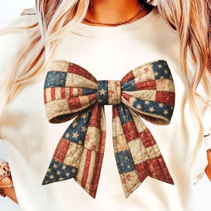 May include: Cream-colored t-shirt featuring a large bow design. The bow is a patchwork of red, white, and blue fabric, resembling the American flag. The bow has a quilted appearance with a detailed pattern, perfect for patriotic events.