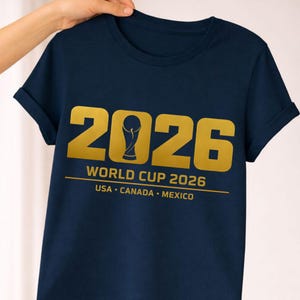 Navy blue shirt mockup displaying a gold Soccer Cup 2026 soccer shirt design. The graphic features bold sports typography showing 2026 with a stylized trophy icon forming the zero and text below reading Soccer Cup 2026 USA Canada Mexico. This soccer themed digital design is perfect for sublimation printing, Cricut and Silhouette cutting machines, print on demand apparel, soccer fan shirts and sports merchandise celebrating the 2026 tournament hosted in North America.