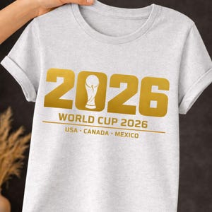 Heather gray shirt mockup displaying a gold North America Global Soccer Cup themed design highlighting the host nations of the 2026 soccer tournament. The design features bold numbers 2026 with a trophy icon in the center and text reading 2026 USA Canada Mexico. This digital sports design is ideal for sublimation printing, Cricut cutting machines, print on demand apparel and custom soccer fan shirts celebrating the international tournament.