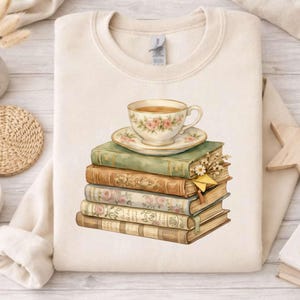 May include: Cream-colored sweatshirt featuring a watercolor design of a teacup and saucer atop a stack of vintage books. The teacup and saucer are decorated with pink roses. The books are in shades of green, brown, and cream.
