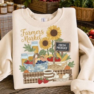 Puede incluir: Sudadera color crema con un diseño de mercado de agricultores. El gráfico incluye girasoles, cestas de productos, tarros y el texto "Fresh Produce" en un letrero. También se representa una taza y fresas.