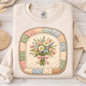 May include: Cream-coloured sweatshirt with a floral bouquet design. The bouquet is tied with a pink bow and set within a quilted, oval frame with pastel patchwork. The sweatshirt has a crew neck and long sleeves.