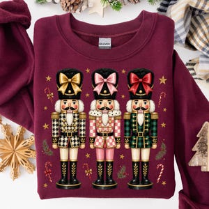 May include: A maroon crewneck sweatshirt featuring a graphic of three nutcrackers standing side by side. The nutcrackers wear gold, pink, and red bows on their hats, with outfits in houndstooth, pink checkered, and green plaid patterns.