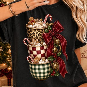 May include: A black t-shirt featuring a graphic of three stacked mugs filled with holiday treats. The top mug has a leopard print, the middle a red and white checkerboard, and the bottom a green and white gingham pattern, adorned with a large red bow.