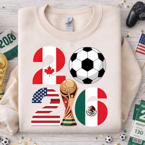 World Soccer Cup 2026 inspired soccer PNG design displayed on a neutral cream sweatshirt mockup, featuring the year 2026 with USA, Canada, and Mexico flag elements, a classic black and white soccer ball, and a golden trophy illustration. This high-resolution transparent PNG is ideal for print on demand shirts, kids soccer apparel, fan merchandise, sublimation projects, and custom football designs celebrating the 2026 world soccer tournament hosted in North America.