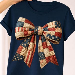May include: Navy blue t-shirt featuring a large bow design. The bow is made of patchwork fabric, incorporating the American flag's stars and stripes in red, white, and blue. The bow is the focal point of the shirt.