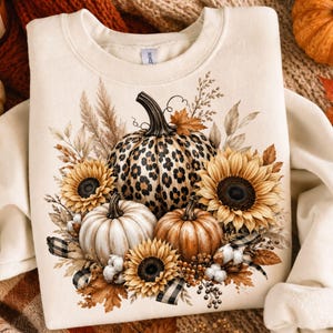 Puede incluir: Una sudadera de cuello redondo color crema con un gráfico central de tres calabazas y girasoles. La calabaza más grande tiene un estampado de leopardo, mientras que las otras son blancas y naranjas, acompañadas de follaje seco y algodón.