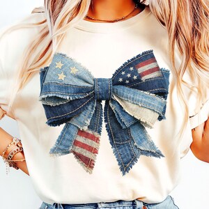May include: A cream-colored t-shirt featuring a large denim bow with a distressed American flag design. The bow is made of layered denim with frayed edges, and includes stars and stripes.