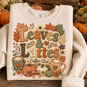 Peut inclure: Sweat-shirt crème avec un motif automnal. Le design affiche les mots "Leaves & Lattes Coffee Club" entourés de feuilles d'automne, de citrouilles et de tasses à café. Les couleurs sont orange, vert et marron.