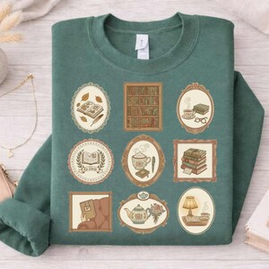 May include: Green sweatshirt with nine framed illustrations of books, teacups, and a teapot. The illustrations are in shades of brown and beige, with details like a stack of books, a cozy reading nook, and a cup of tea.