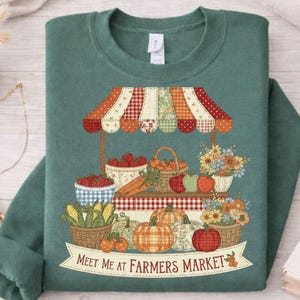 May include: Green sweatshirt featuring a farmers market design. The graphic shows a market stall with fruit, vegetables, and flowers. The text "Meet Me at Farmers Market" is displayed on a banner.