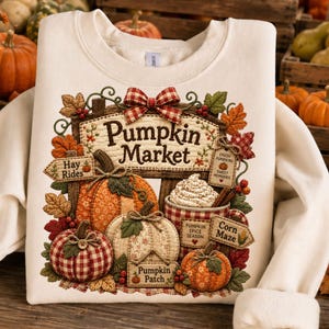 May include: A cream-colored sweatshirt featuring a graphic of a pumpkin market with pumpkins, a mug of whipped cream, and signs reading Pumpkin Market, Hay Rides, Fresh Pumpkins, Sweet Memories, Pumpkin Spice Season, Corn Maze, and Pumpkin Patch.